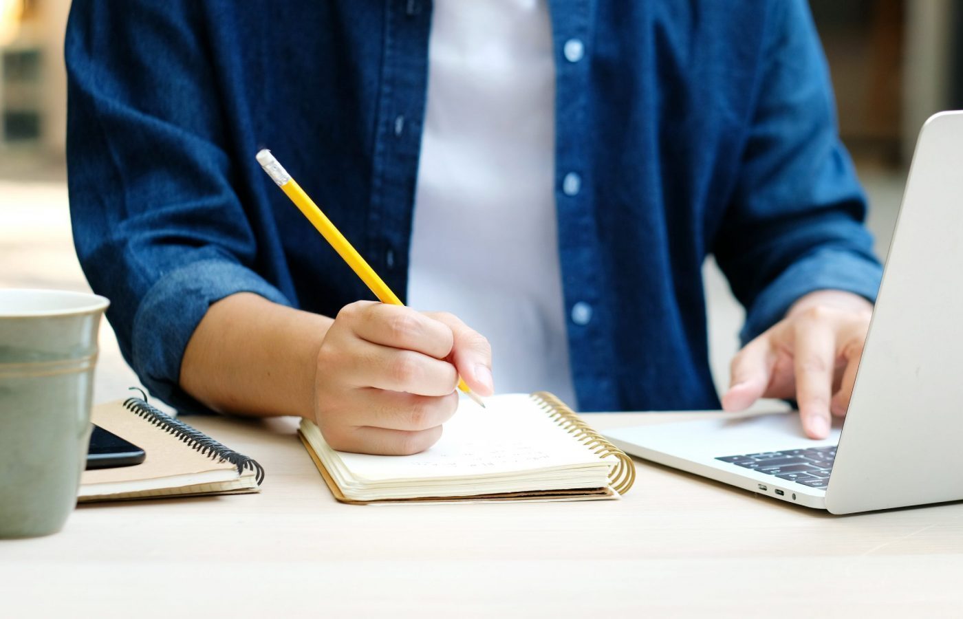 Student writing on notebook while using a laptop